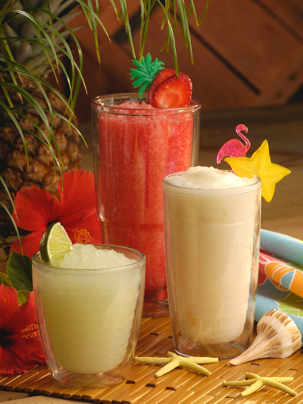 Image shows three summer blended drinks with fruit garnishes in a beachy setting. 